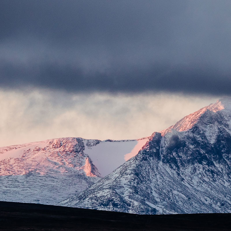 A snow-covered mountain beneath dark clouds, glowing in the light of the sunset.