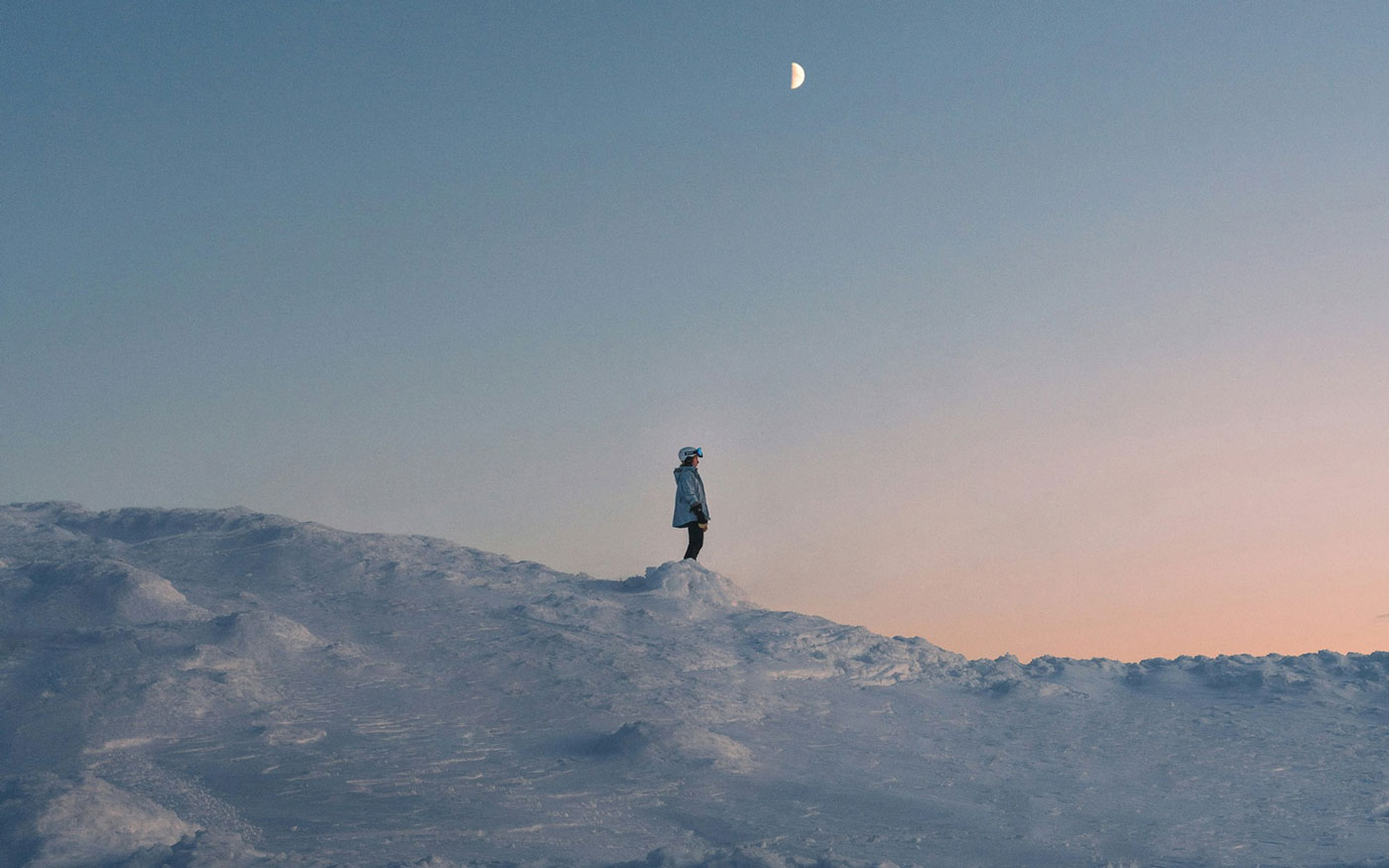 A female skier stands on a snow-covered ridge at sunset, gazing out over the snowy mountains.