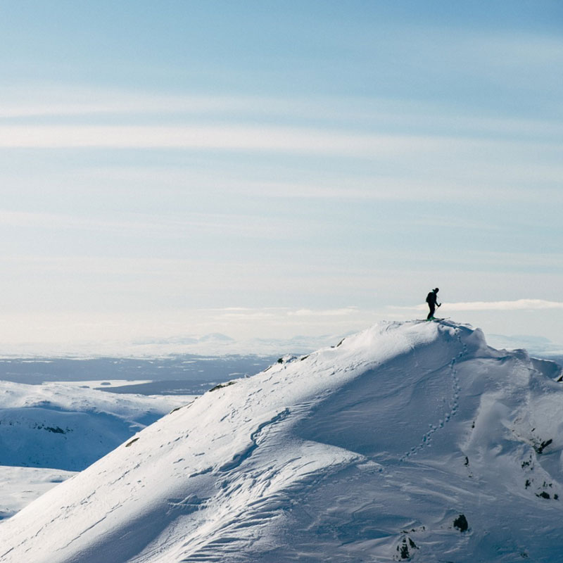 Skier atop a snow-covered peak under a sunny sky, overlooking a winter landscape.